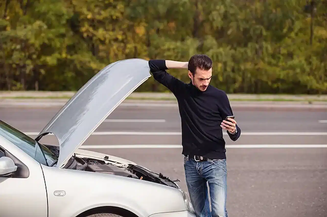 Driver standing beside a broken down car with the hood open while checking phone for roadside assistance