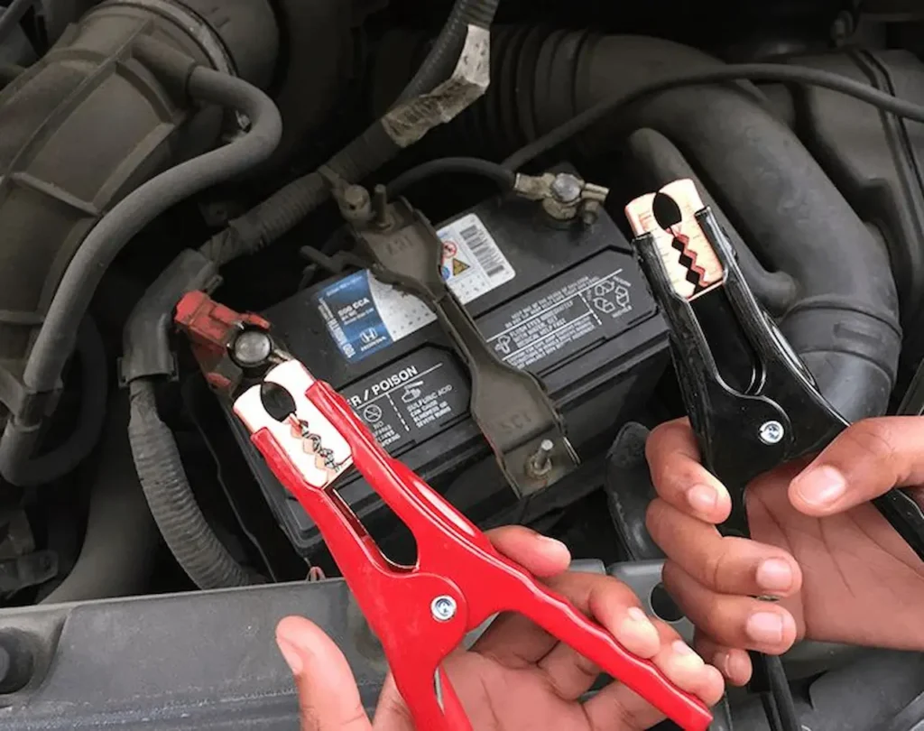 Hands holding red and black jumper cable clamps near a car battery, preparing for a battery jump start.