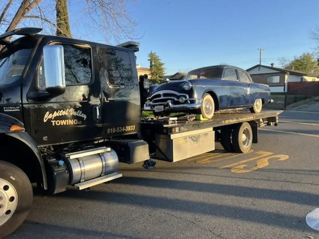 Tow truck transporting a classic car on a flatbed in a residential area.