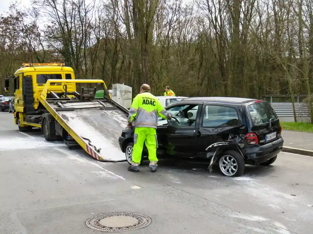 A roadside assistance worker in a high-visibility jacket preparing a yellow flatbed truck to tow a crashed black car.